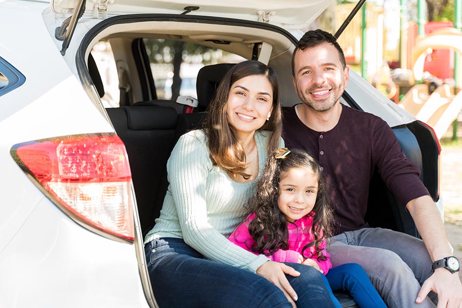 Personal Insurance - Mother, Father and Young Daughter Sit In the Back Hatch of Their Silver Car, Smiling